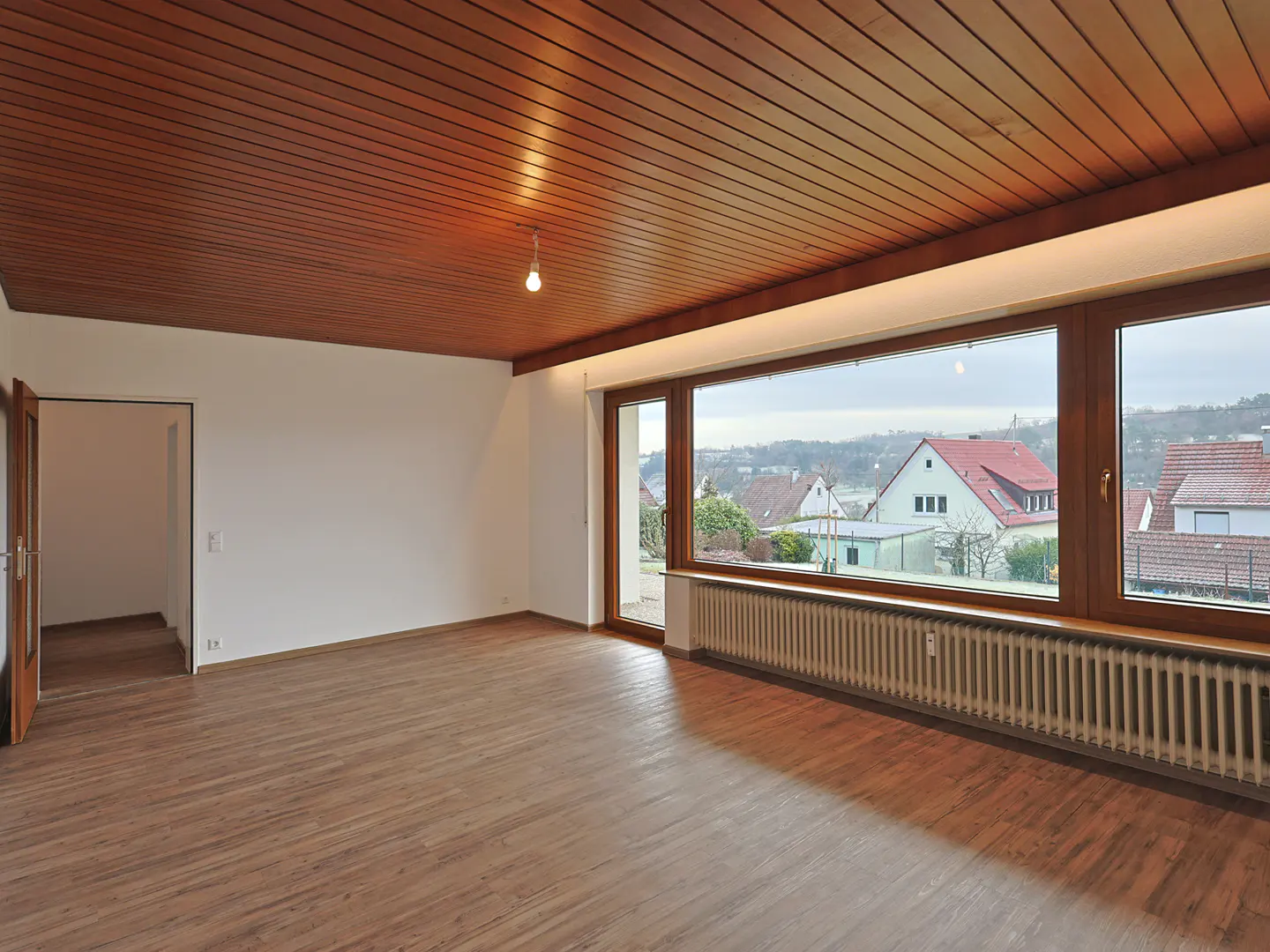 Empty room with wood-paneled ceiling, light wood floors, and large windows overlooking houses.