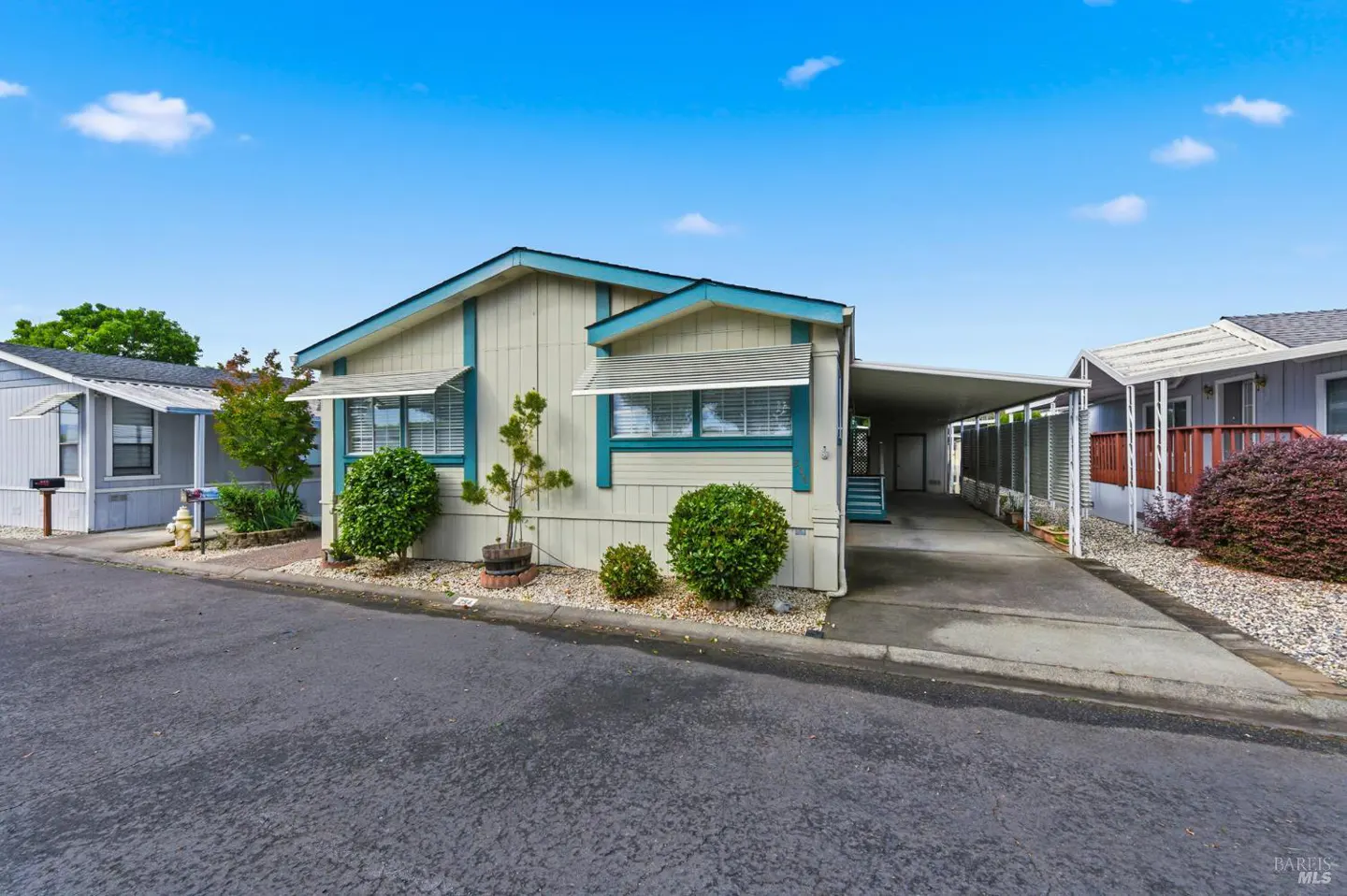 A light-yellow mobile home with blue trim and a carport on a sunny day.