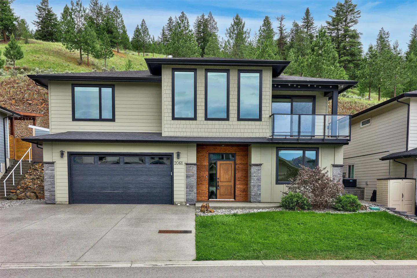 Modern two-story home with sage green siding, dark garage, wood-paneled entryway, and a glass balcony against a forest.