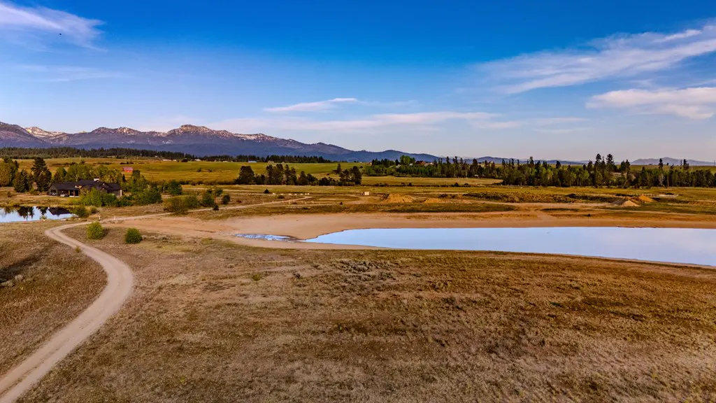 Aerial view of a dirt path winding through a dry, grassy landscape toward a pond with snow-capped mountains in the distance.