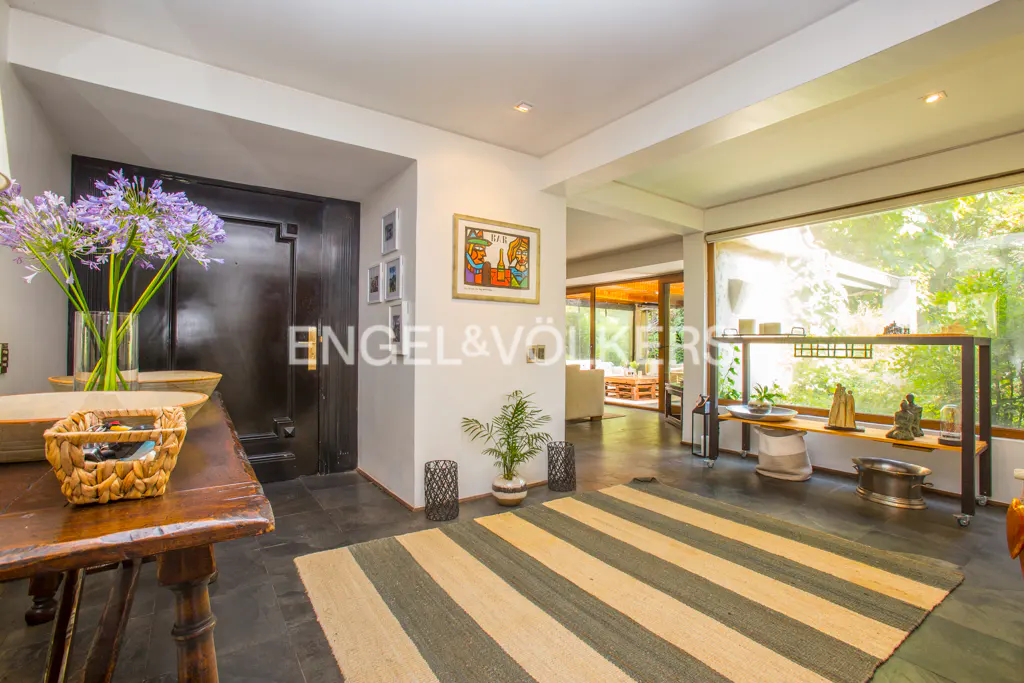 A home's entryway with a dark door, wooden table with flowers, and a striped rug leading to a bright living area.