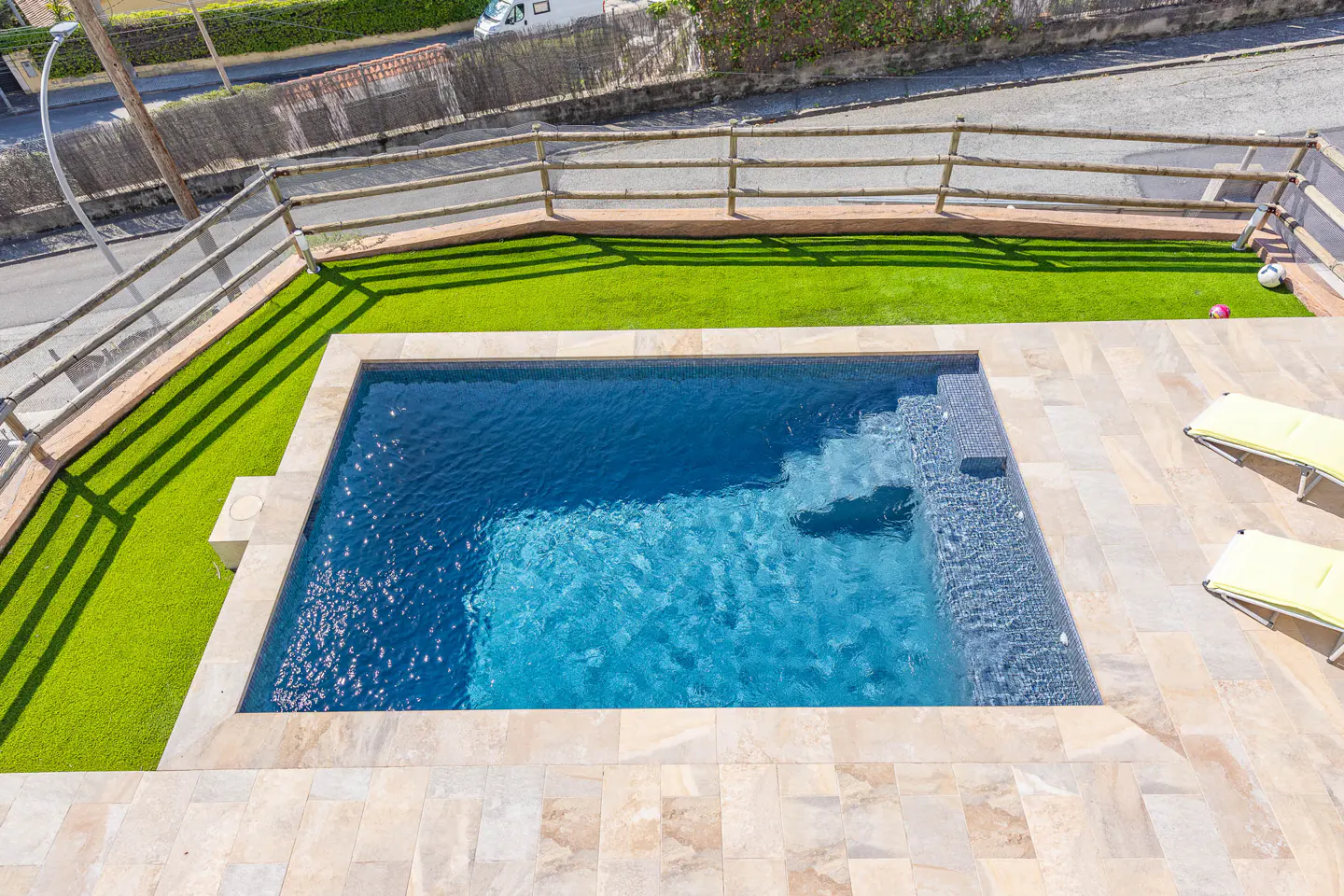 Aerial view of a rectangular pool with blue water, surrounded by a stone patio, green grass, and a wooden fence. Two yellow lounge chairs are on the patio.