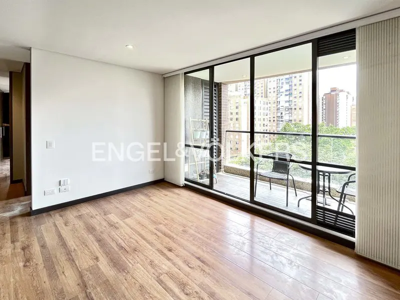 Bright, empty room with wood floors and white walls. A balcony with chairs and a table overlooks a city with green trees.