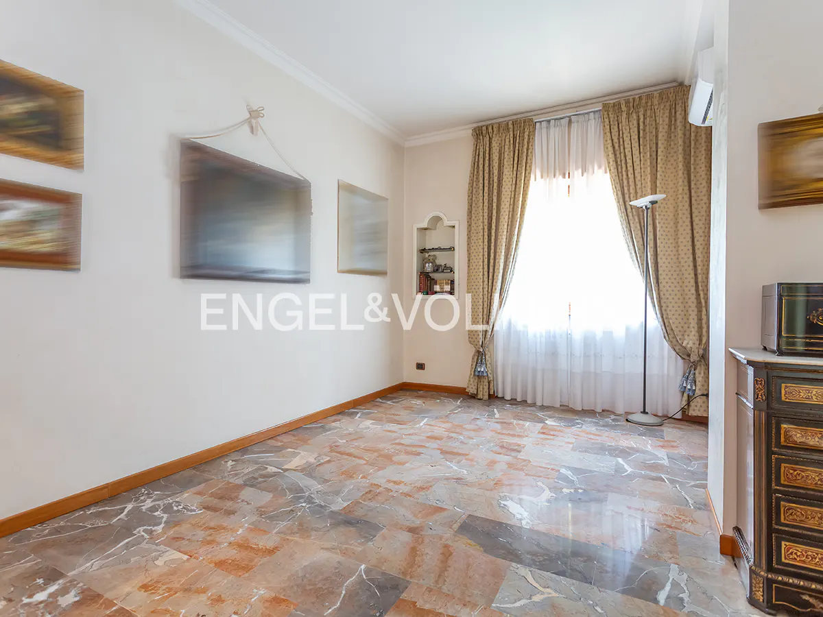 A bright room with marble floors, beige walls, and gold curtains framing a window. Artwork hangs on the wall.