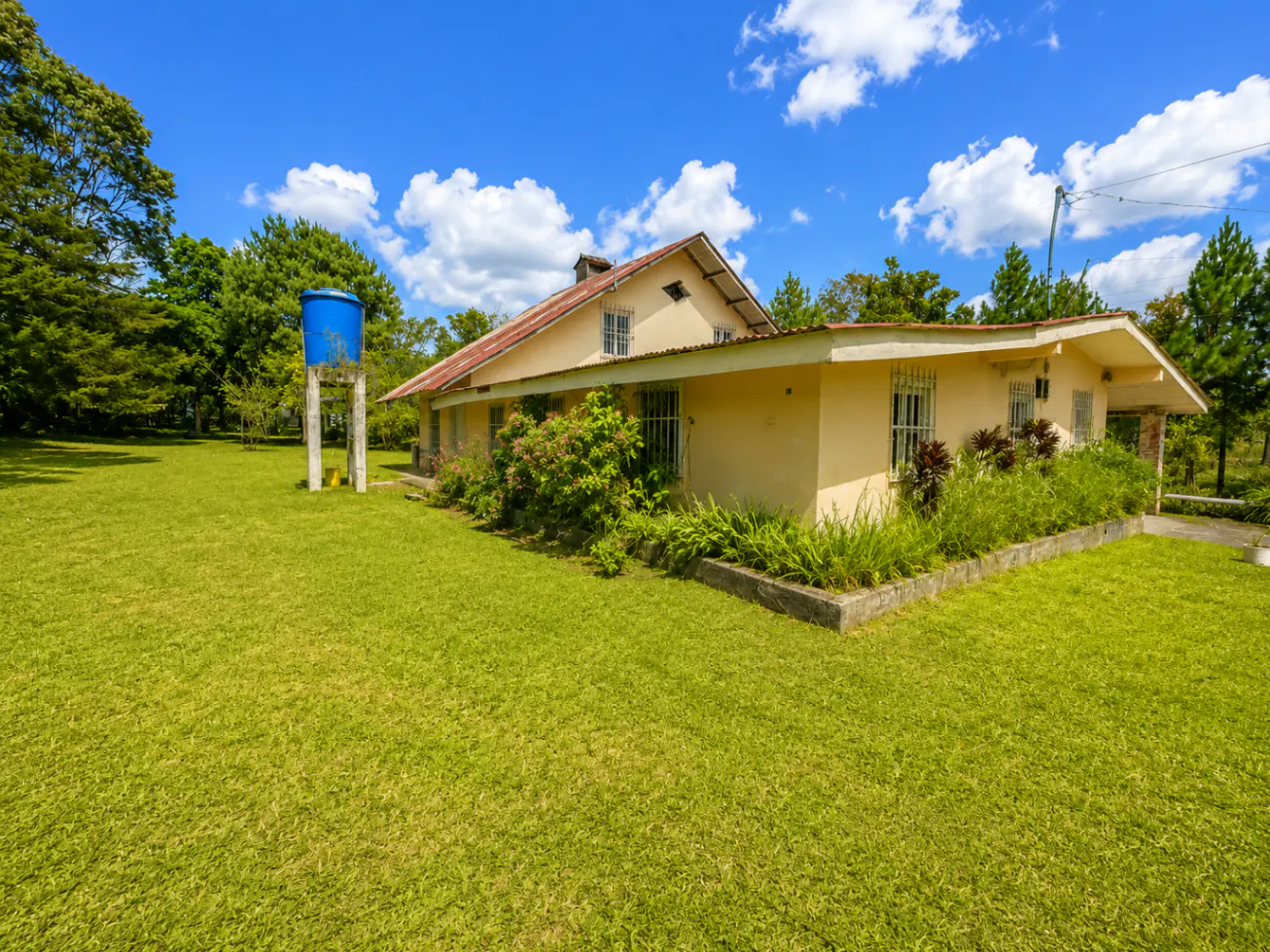 Tan house with a red roof and barred windows, surrounded by a green lawn and trees under a blue sky with white clouds. A blue water tank stands nearby.