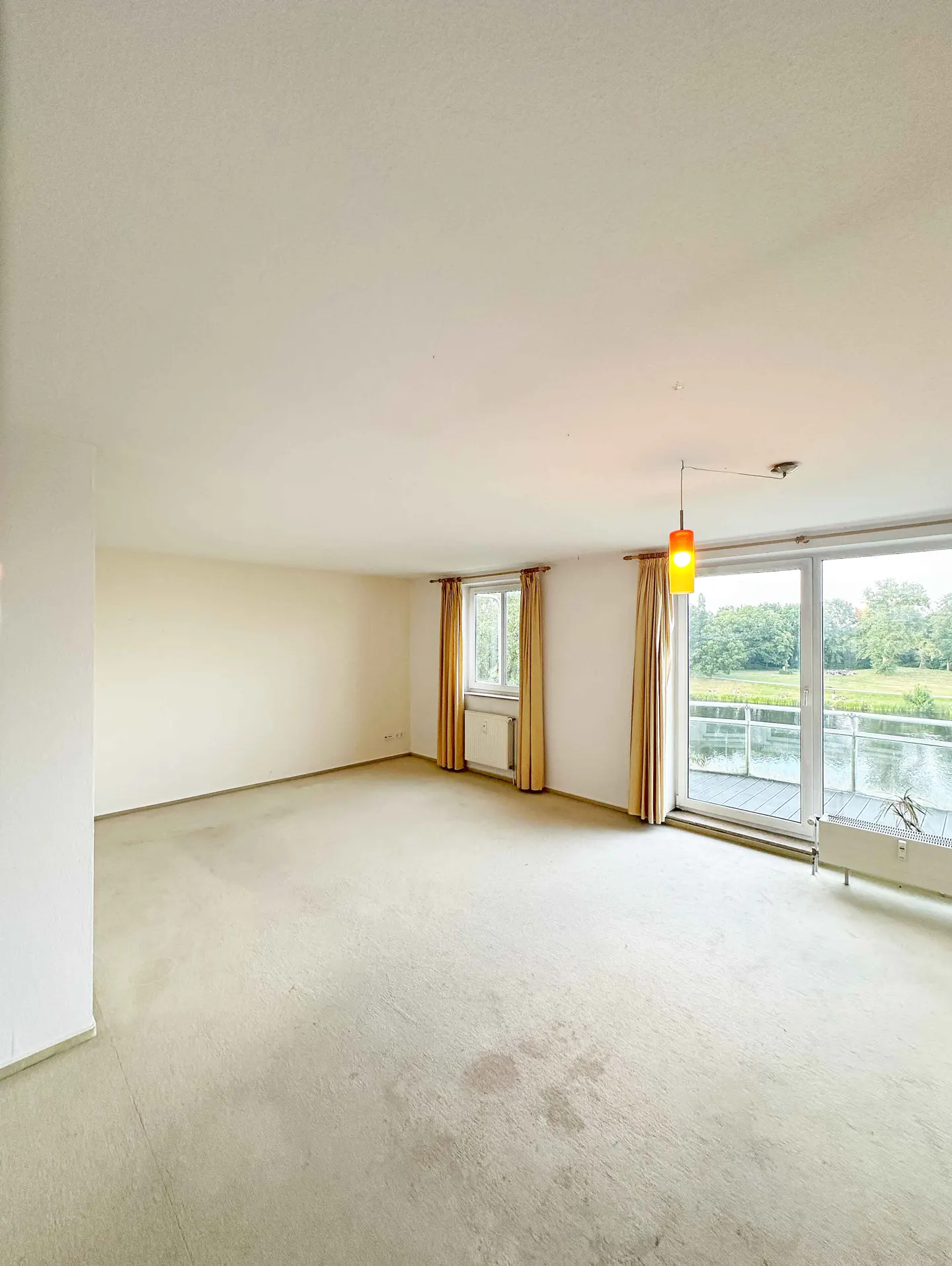Spacious, empty room with beige carpet, white walls, tan curtains, and a large window overlooking a river and greenery.