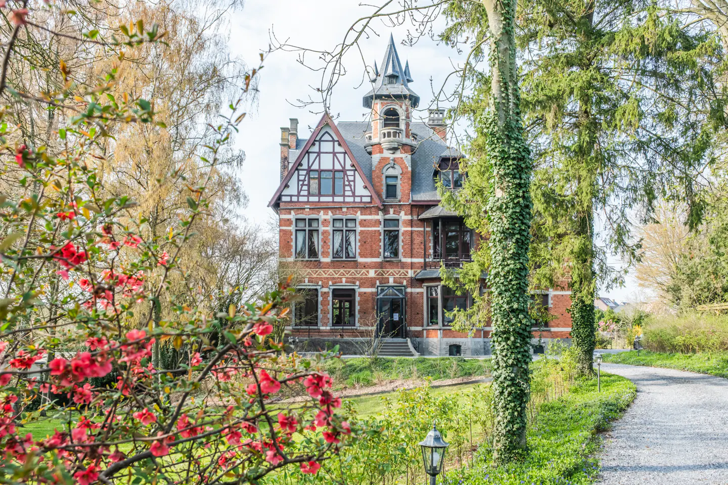 A three-story brick mansion with a tower, surrounded by trees and flowering bushes. A gravel path leads to the house.