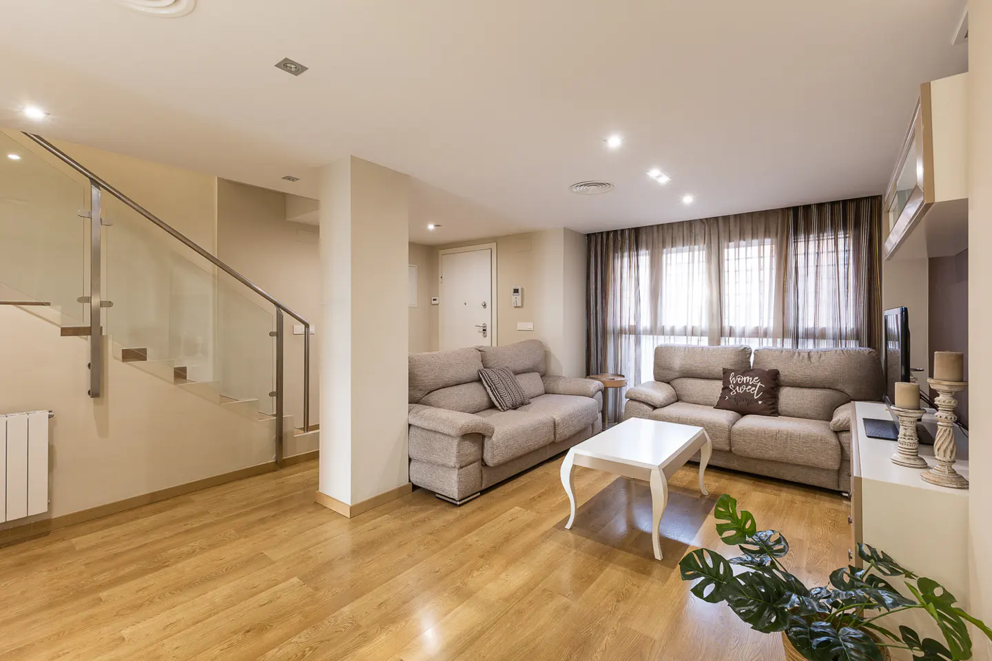 A bright living room with wood floors, beige sofas, a white coffee table, and a staircase with a glass railing.