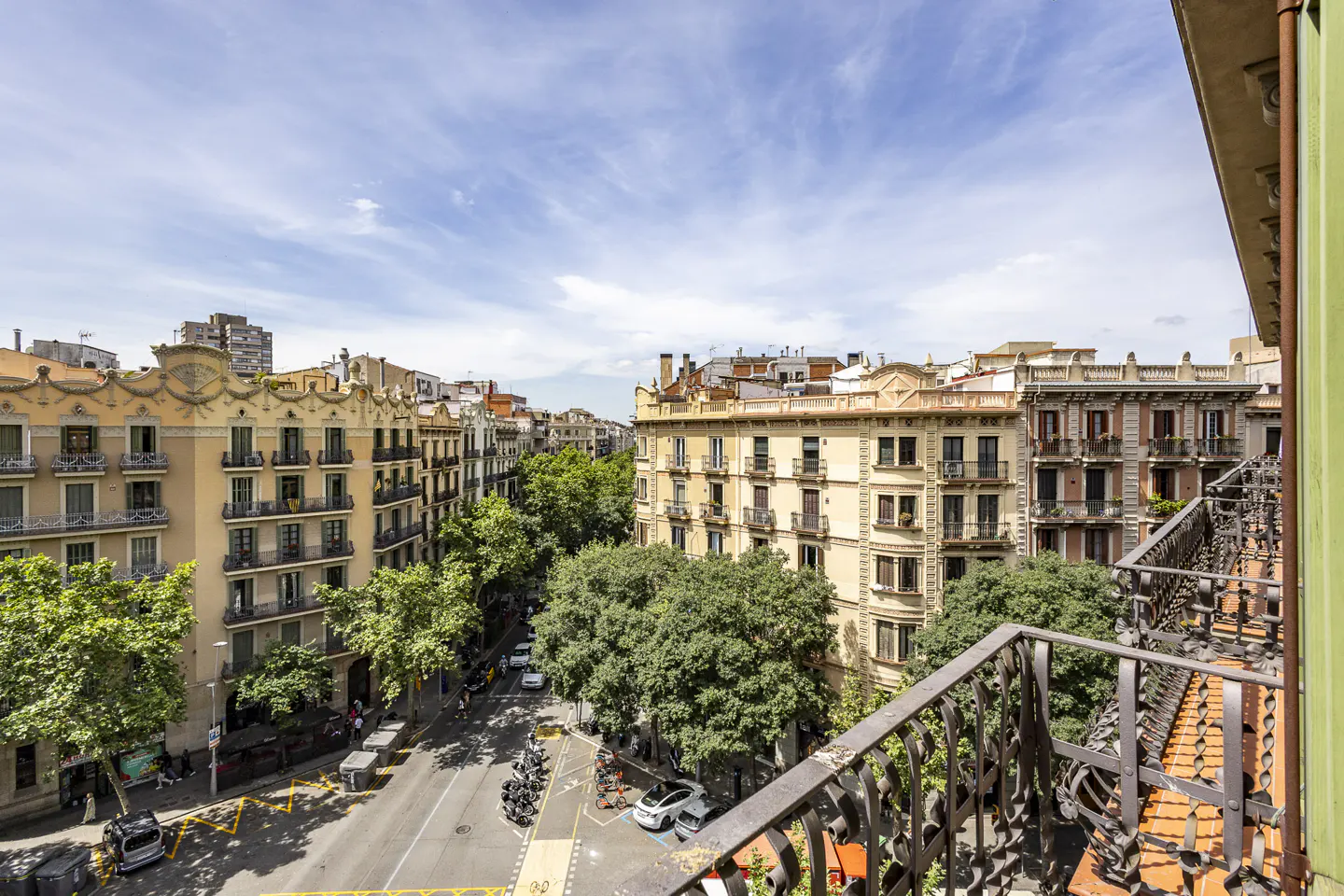 View from a balcony overlooking a sunny Barcelona street with historic tan buildings, lush green trees, and parked cars.
