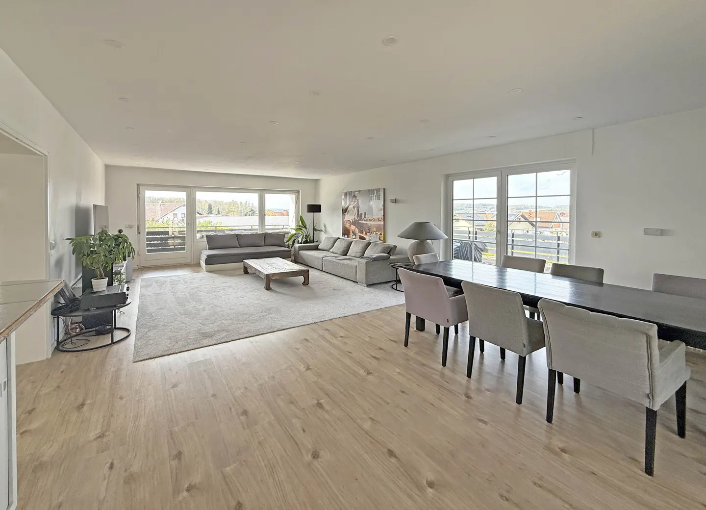 Bright, open-plan living and dining area with wood floors, grey sofas, and a long dark wood table with upholstered chairs. Large windows offer outdoor views.