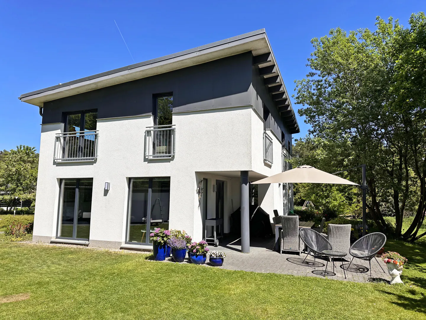 Modern two-story white and grey house with a patio, outdoor furniture, umbrella, and a lush green lawn under a blue sky.