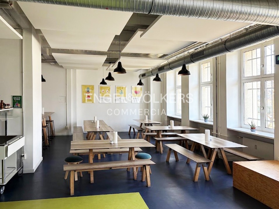 Modern office cafeteria with wooden tables, benches, and stools under industrial-style ceiling lights and exposed ductwork.