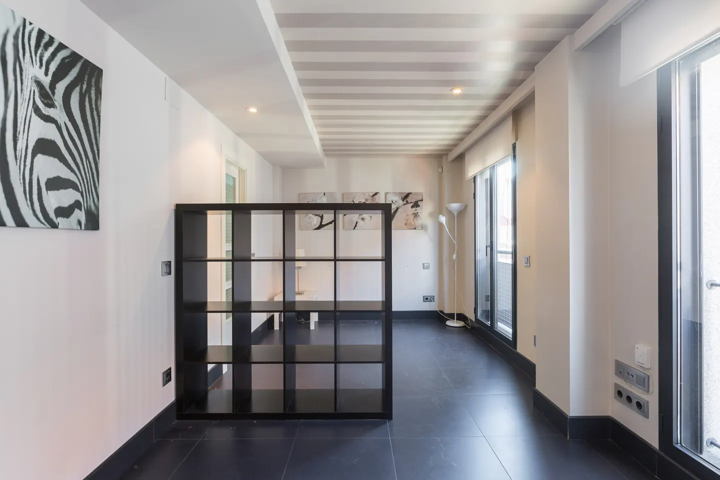 A modern apartment hallway with black tile floors, white walls, and a black shelving unit. A zebra print hangs on the wall.