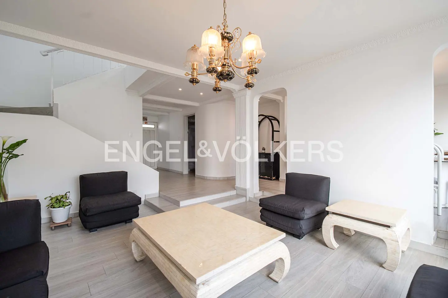 Bright living room with white walls, gray floors, and black chairs. A large, light-colored coffee table sits in the center, under a decorative chandelier. Stairs lead to another level.
