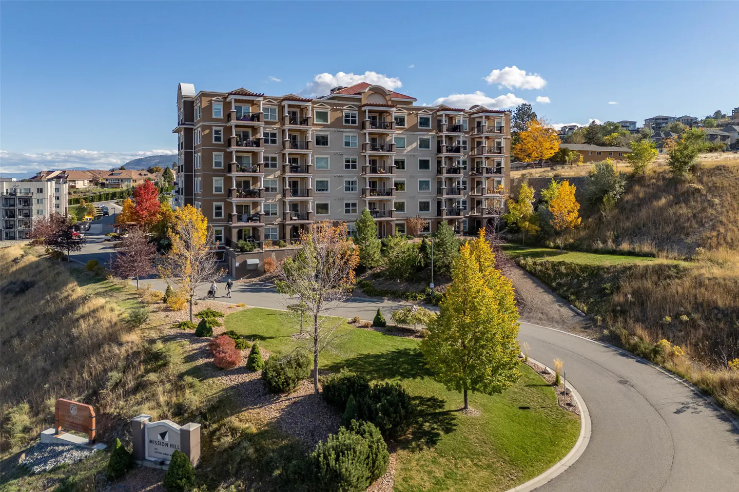 Exterior view of a multi-story condo building with balconies, set on a grassy hill with autumn trees and a winding road.