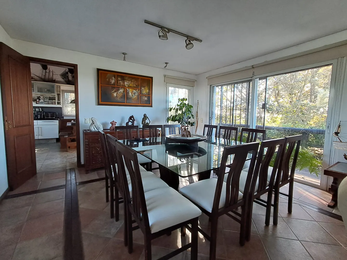 Bright dining room with a large glass table, dark wood chairs with white cushions, and a view of trees through sliding glass doors.
