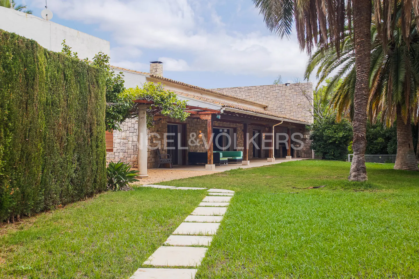 Stone house with a covered patio, green lawn, and a stone path leading to the house. Palm trees are on the right.