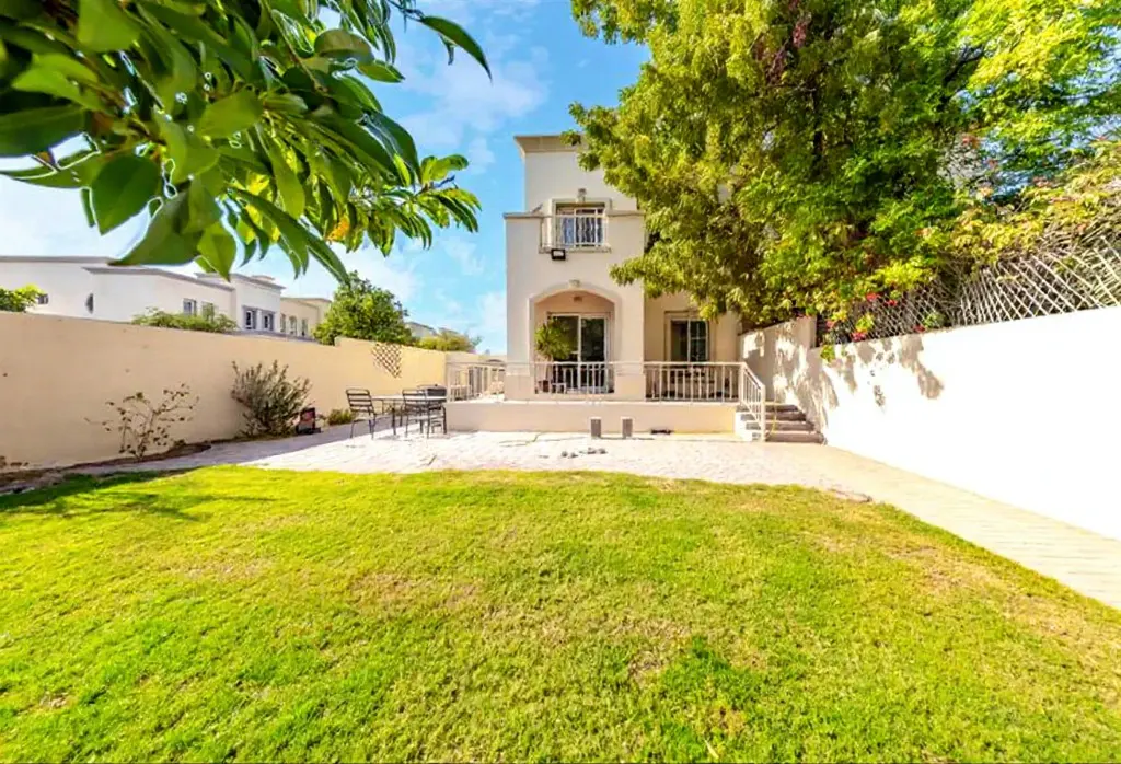 A two-story white house with a balcony overlooks a green lawn and patio with outdoor furniture.