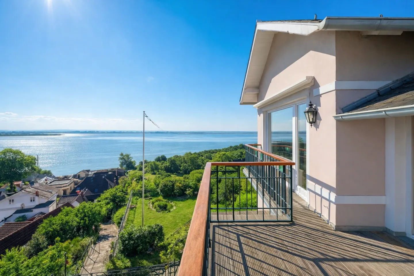 View from a wood-decked balcony with black railings, overlooking green trees and a blue ocean under a clear sky.
