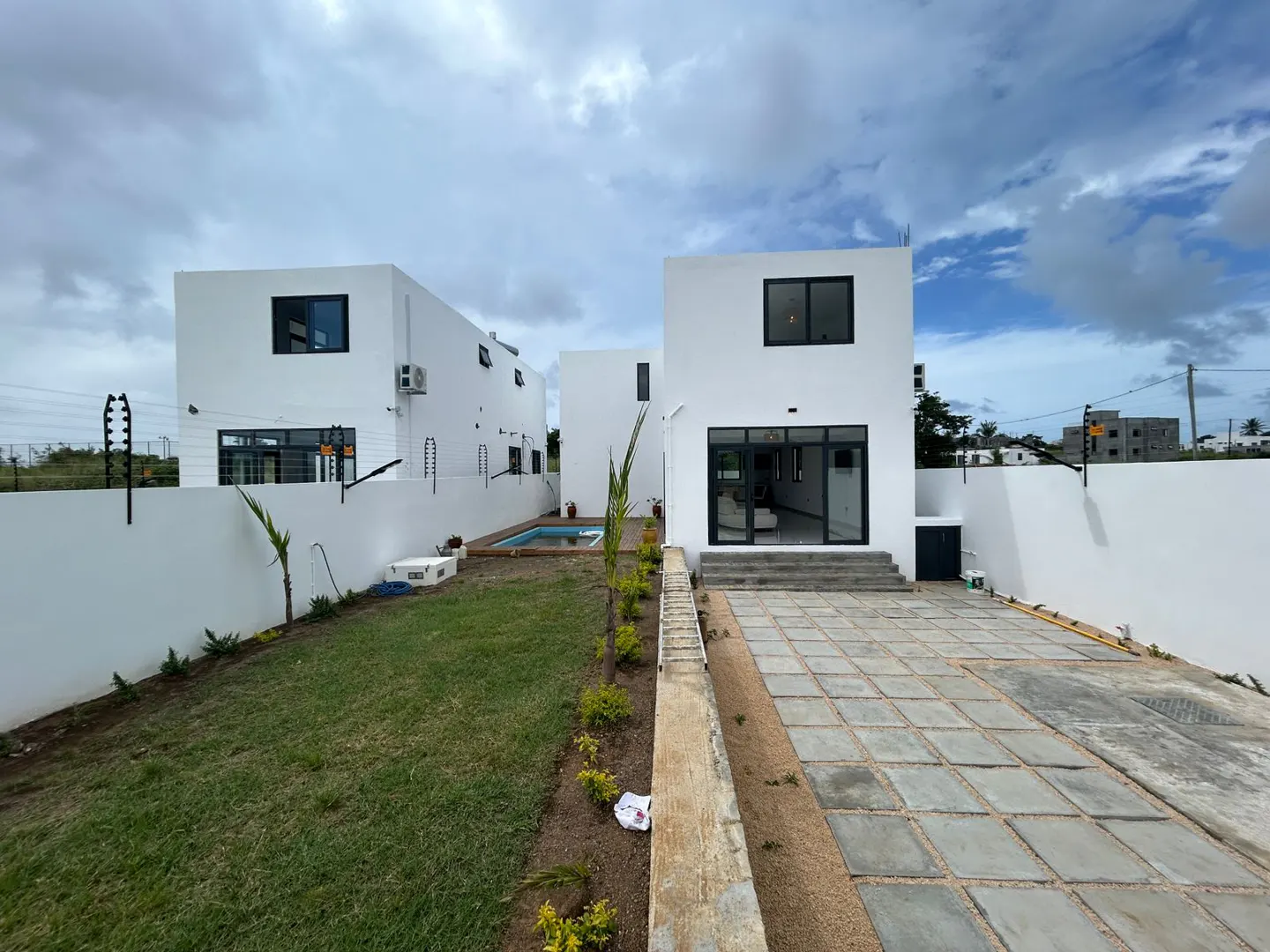 Modern white houses with black framed windows, a small pool, and a stone-paved patio under a cloudy sky.