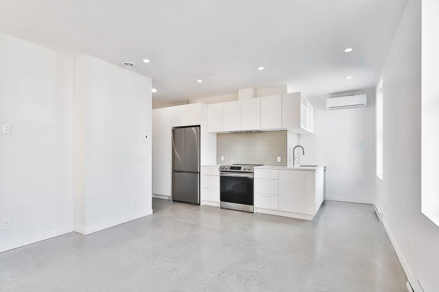 Bright, modern apartment with white walls, grey concrete floors, and stainless steel appliances in the kitchen area.