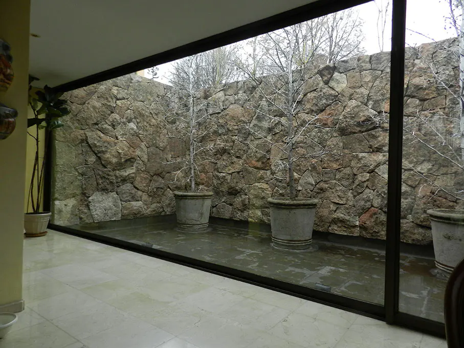 Indoor courtyard with stone wall, potted trees, and large glass windows. The floor is light-colored tile.