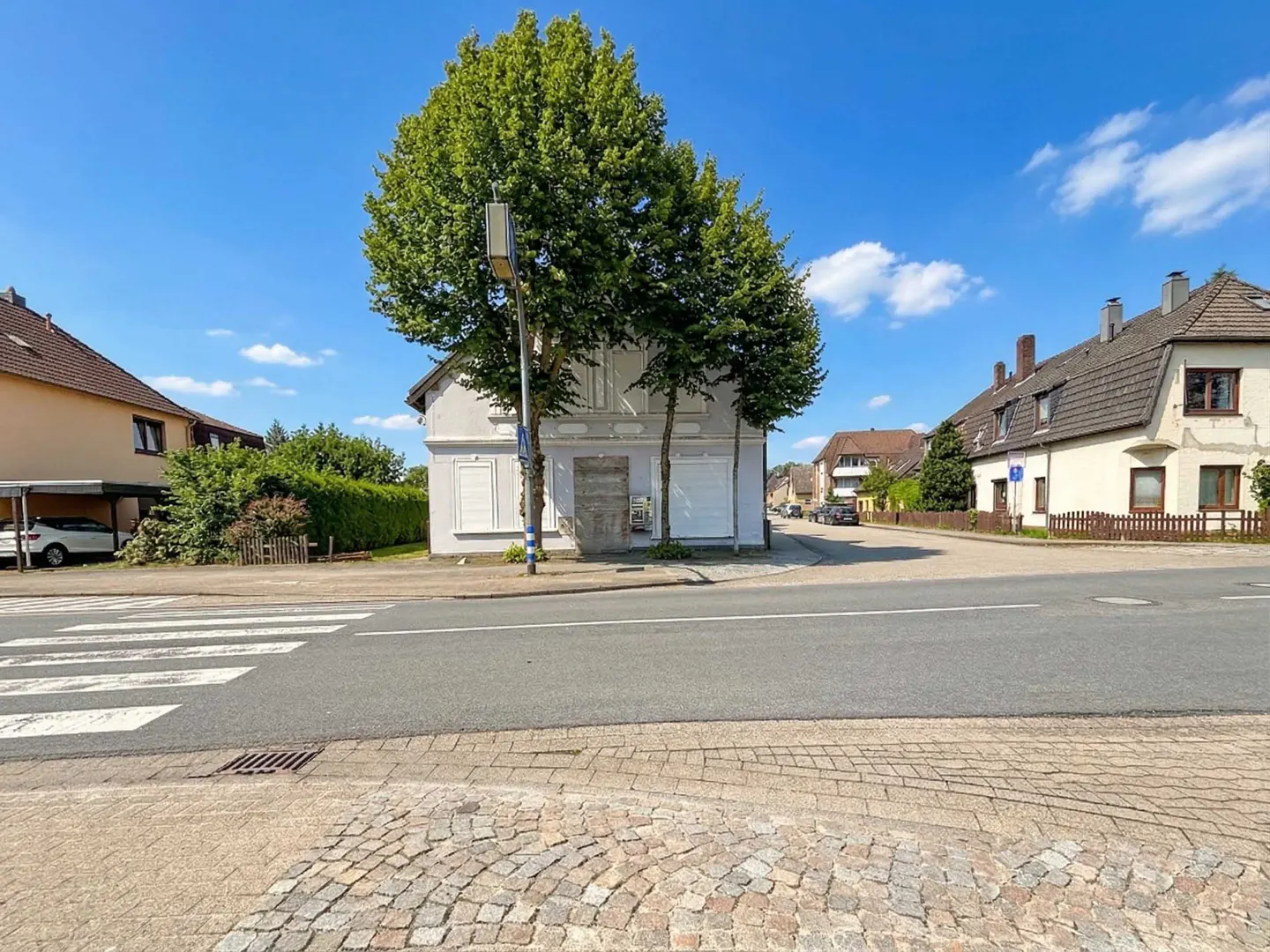 Street view of a two-story white building with boarded-up windows, framed by green trees under a blue sky.