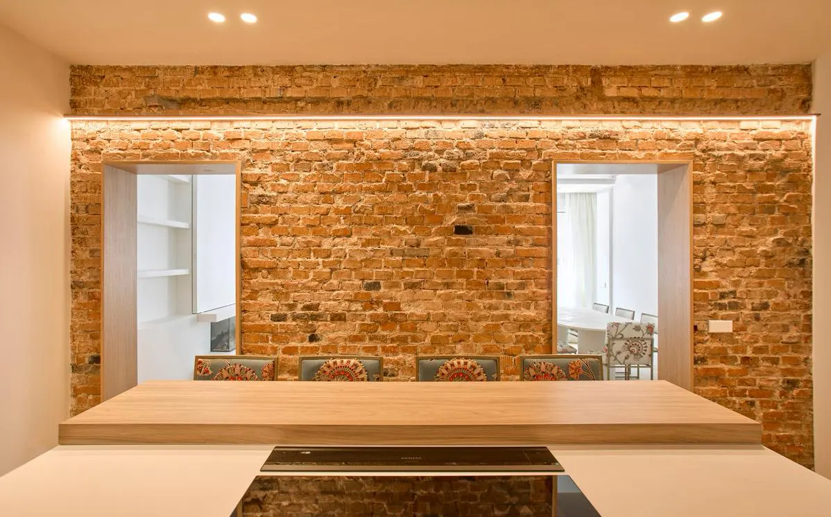 Interior view of a kitchen with a brick wall, two doorways, a wooden countertop, and chairs with patterned upholstery.