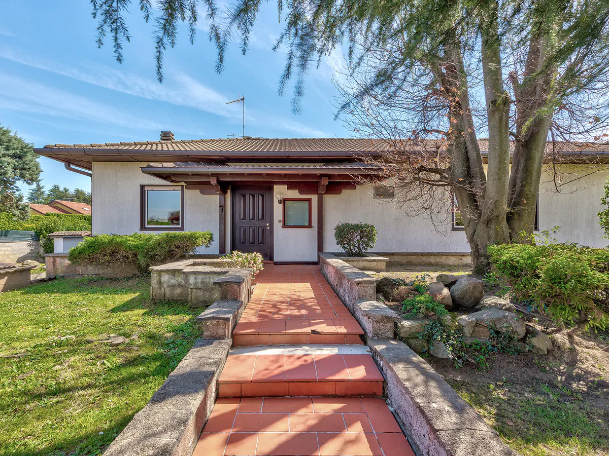 Front exterior of a one-story white house with a brown roof and a red tile walkway. A tree is on the right.