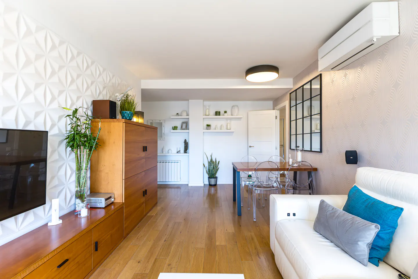 Bright living room with wood floors, a white sofa with blue pillows, and a wooden cabinet against a geometric patterned wall.