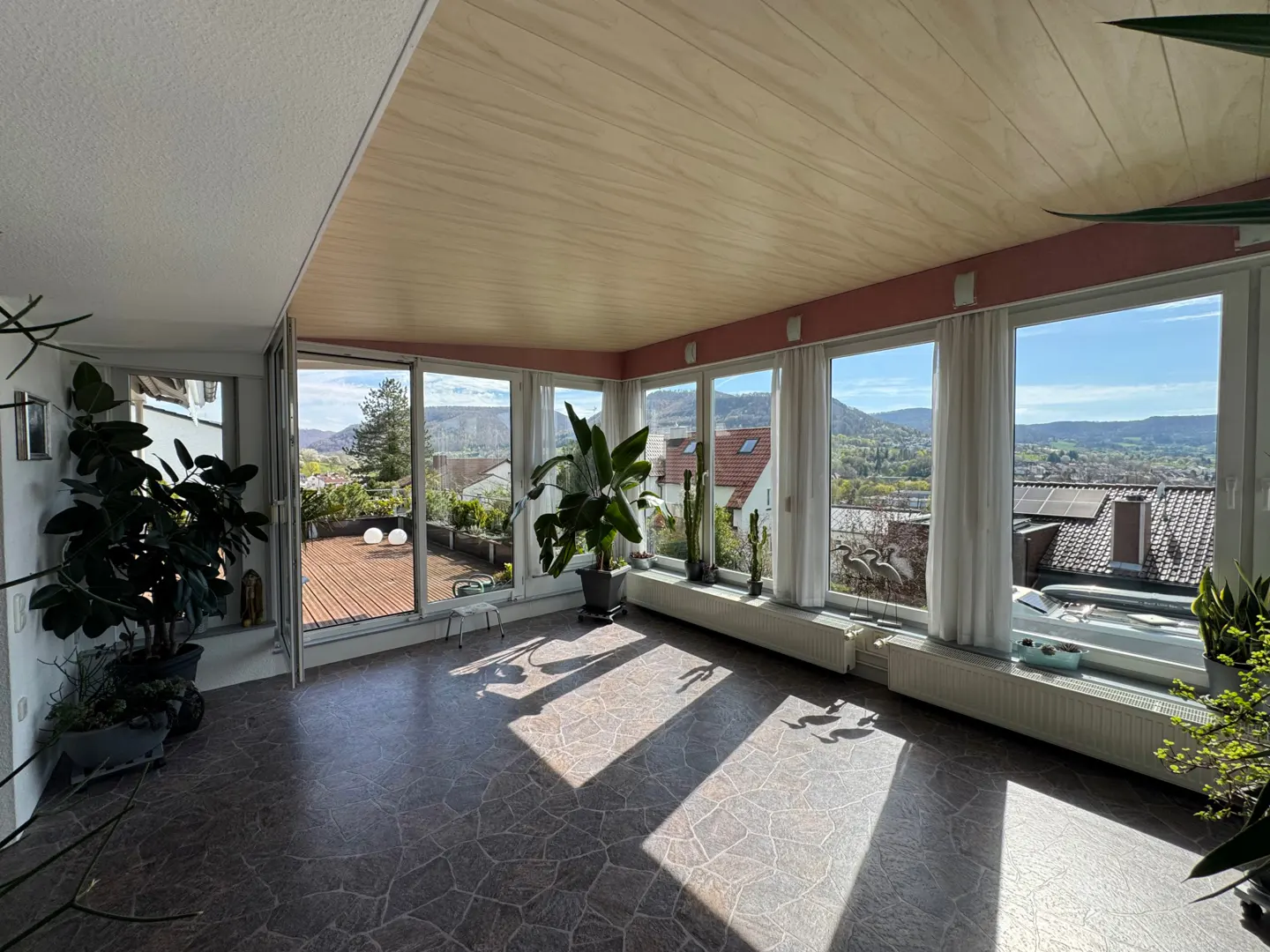 Bright sunroom with large windows, plants, and a view of a town and hills. The floor is brown tile, and the ceiling is light wood.