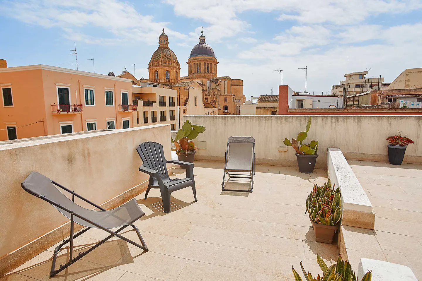 Rooftop view with lounge chairs, potted plants, and a city view. A church with domes is visible in the background.