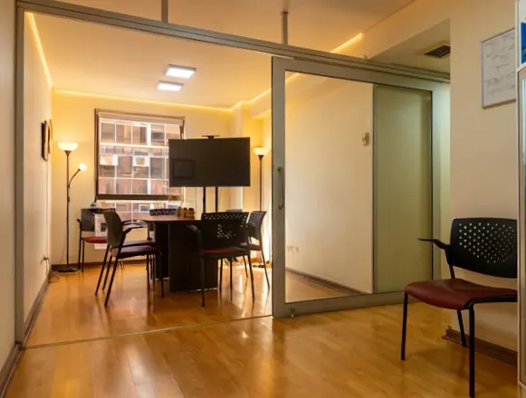 Office space with a conference room visible through a sliding glass door. The room has a table, chairs, and a large screen.