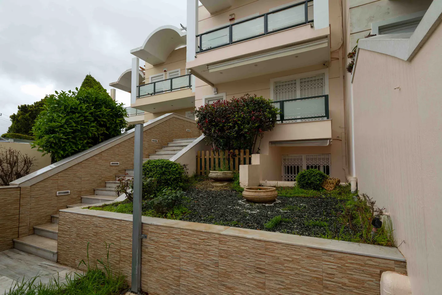 Exterior view of a multi-story beige building with balconies, stairs, and landscaping on a cloudy day.