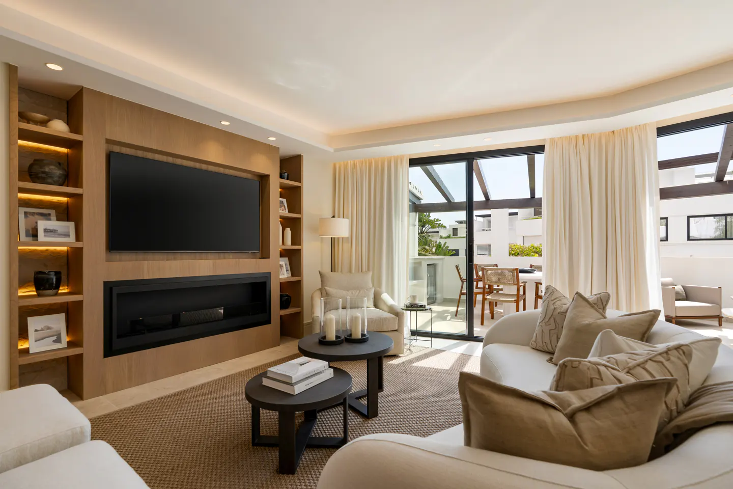 Bright living room with a large TV above a fireplace, bookshelves, white sofas, and a balcony with outdoor seating.