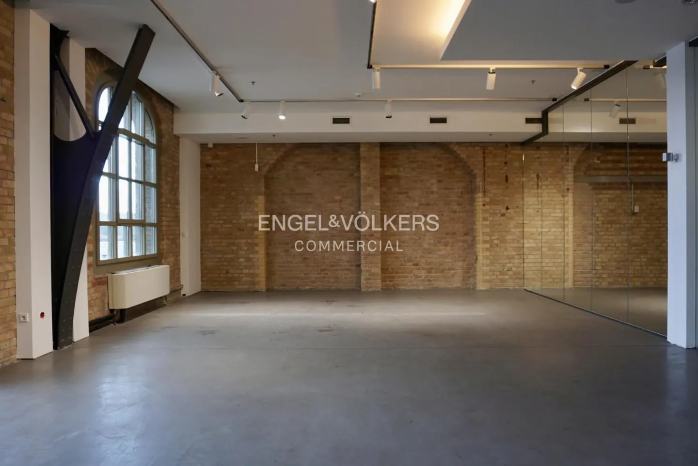 Empty commercial space with brick walls, concrete floor, and large windows. Engel & Völkers logo on the wall.
