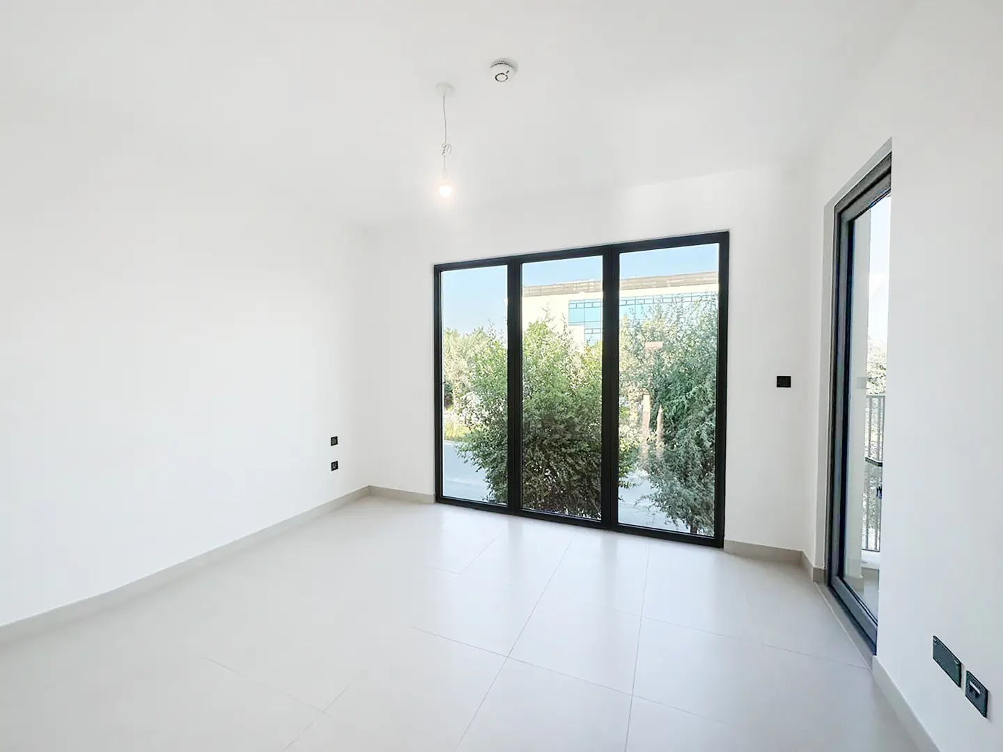 Bright, empty room with white walls and tile floor. Large black-framed windows overlook green trees and a building.