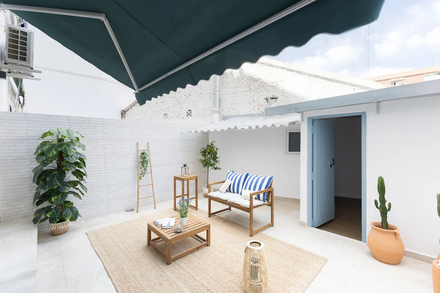 Outdoor patio with a wooden bench, table, and rug. A blue awning provides shade. White brick walls and potted plants add to the ambiance.