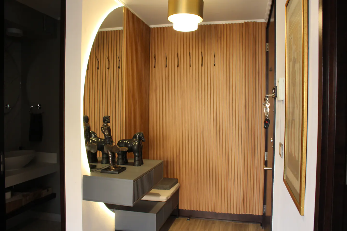 Entryway with wood-paneled wall, coat hooks, and a gray console table with sculptures. A round mirror and a bathroom are visible.