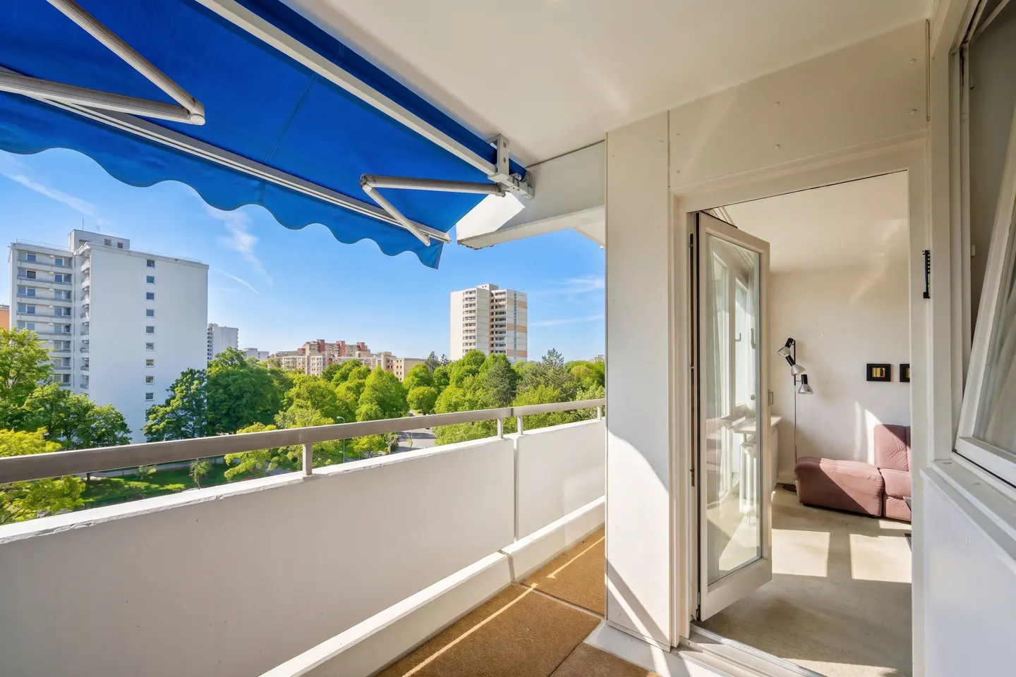 Balcony view with blue awning, white railing, and open door to a bright living room with a brown sofa. Cityscape background.