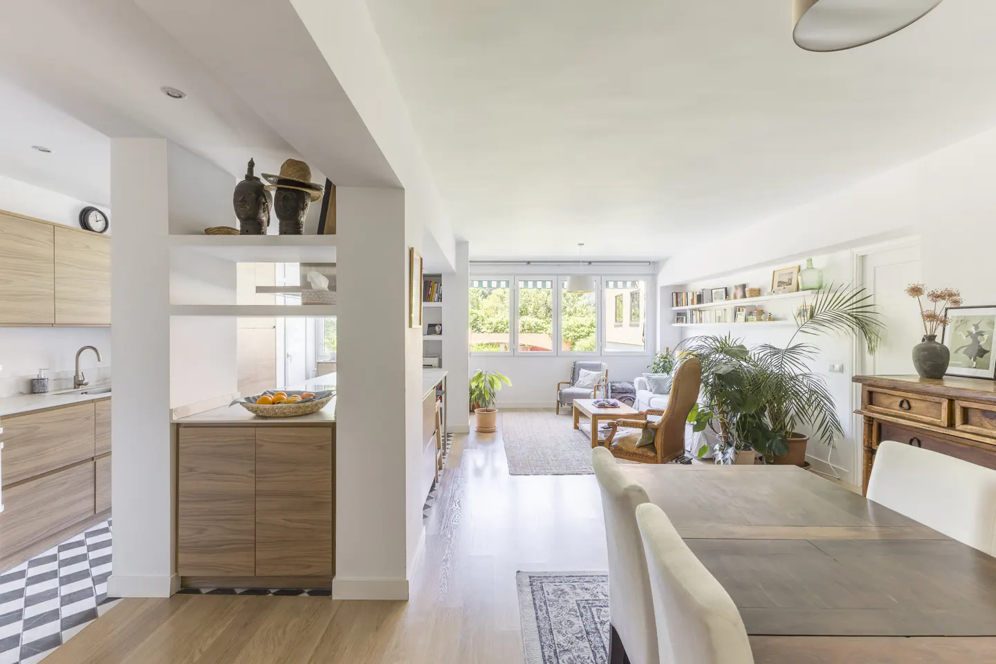 Bright, open-concept living space with wood floors, white walls, and large windows. Kitchen with light wood cabinets and black-and-white tile floor. Dining table with white chairs.