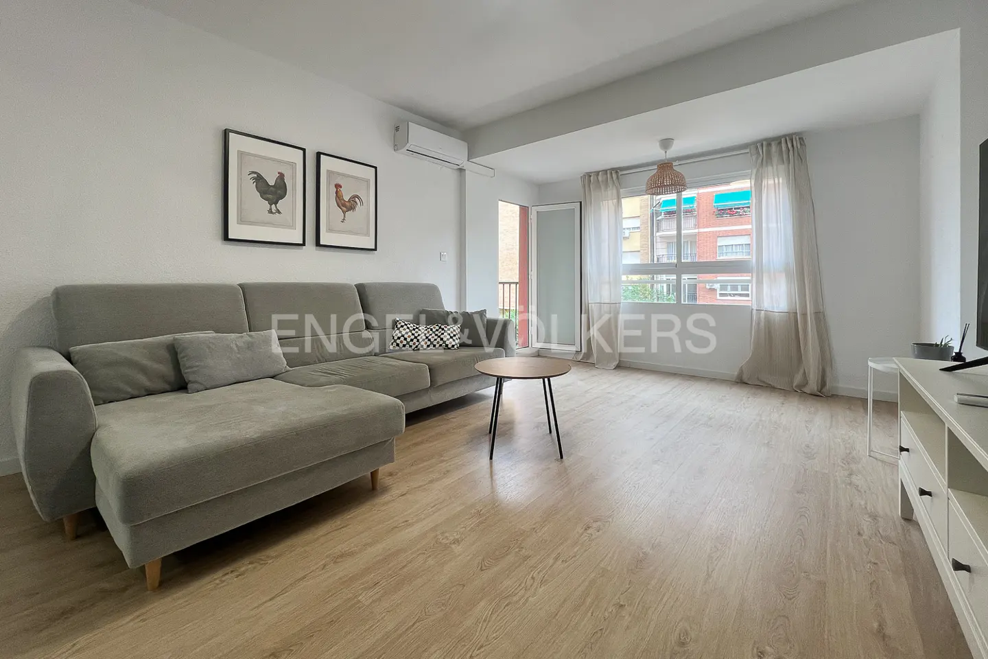 Living room with a gray sectional sofa, wood floors, and two framed rooster prints on the wall. A round coffee table sits in front of the sofa.