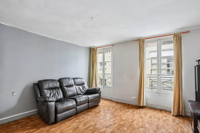 Living room with a black leather sofa, wood floors, and two windows with beige curtains.