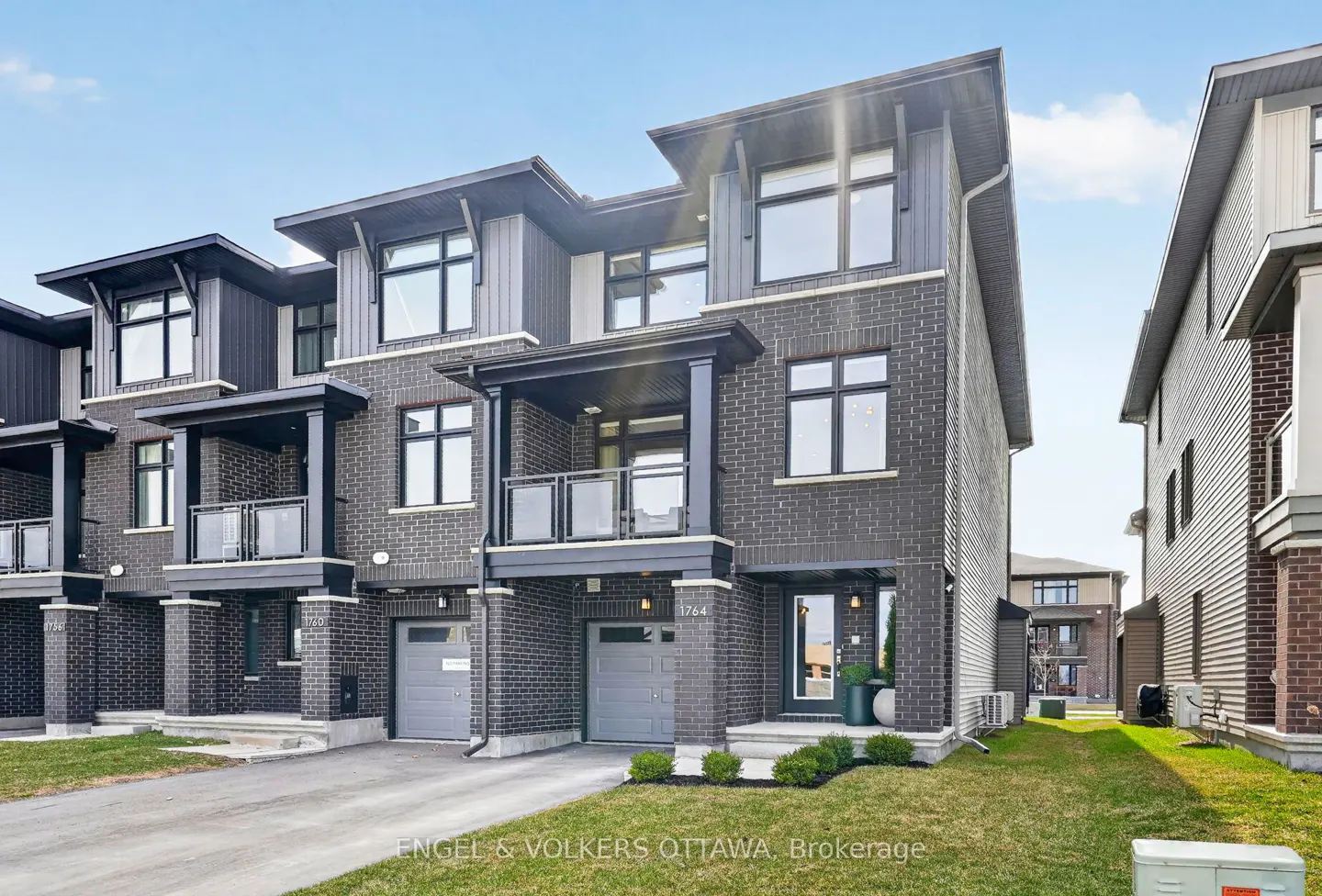 Exterior of modern, three-story townhouses with gray brick, black trim, and small balconies under a blue sky.