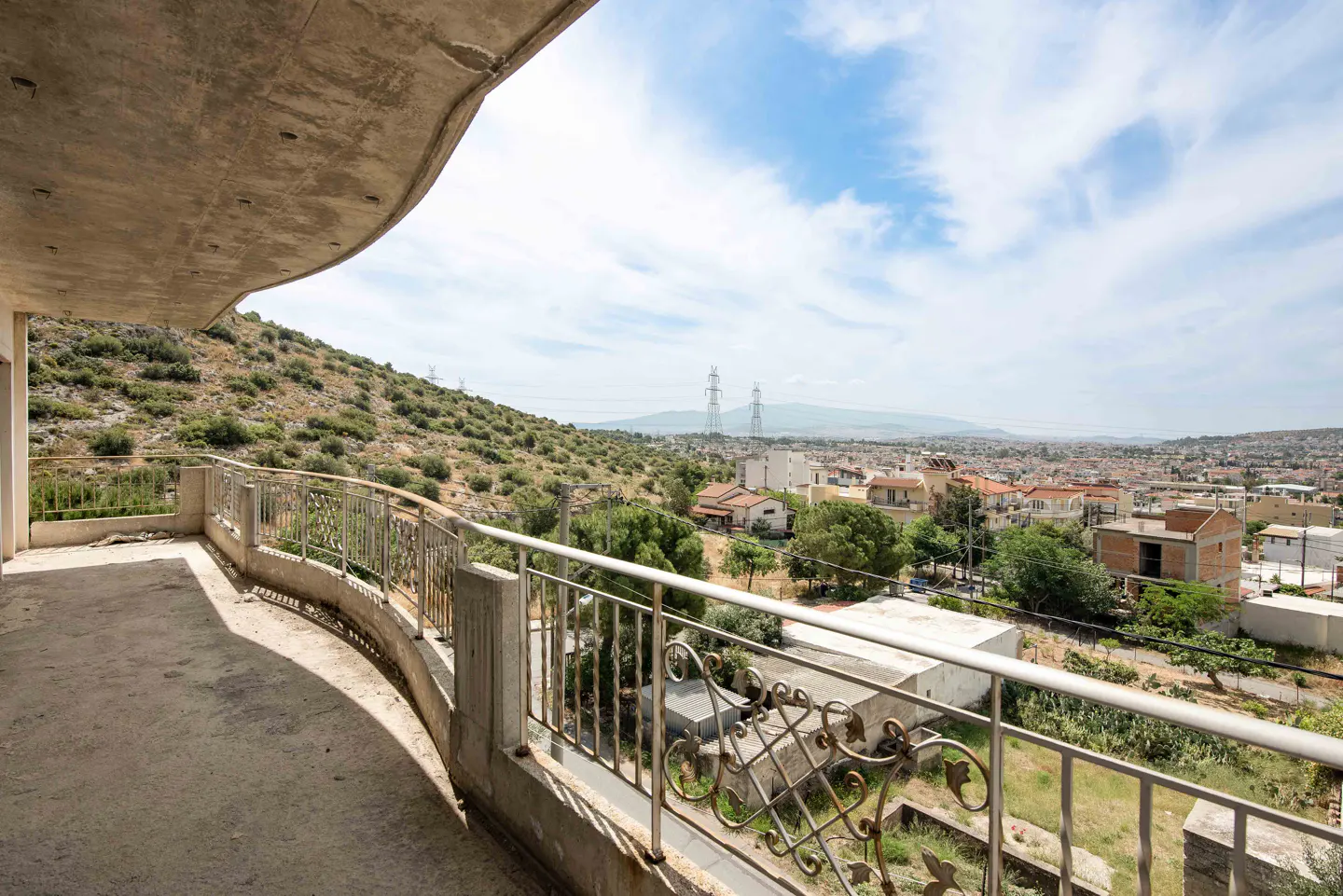 Curved concrete balcony with metal railing overlooking a hillside, town buildings, and distant mountains under a blue sky.