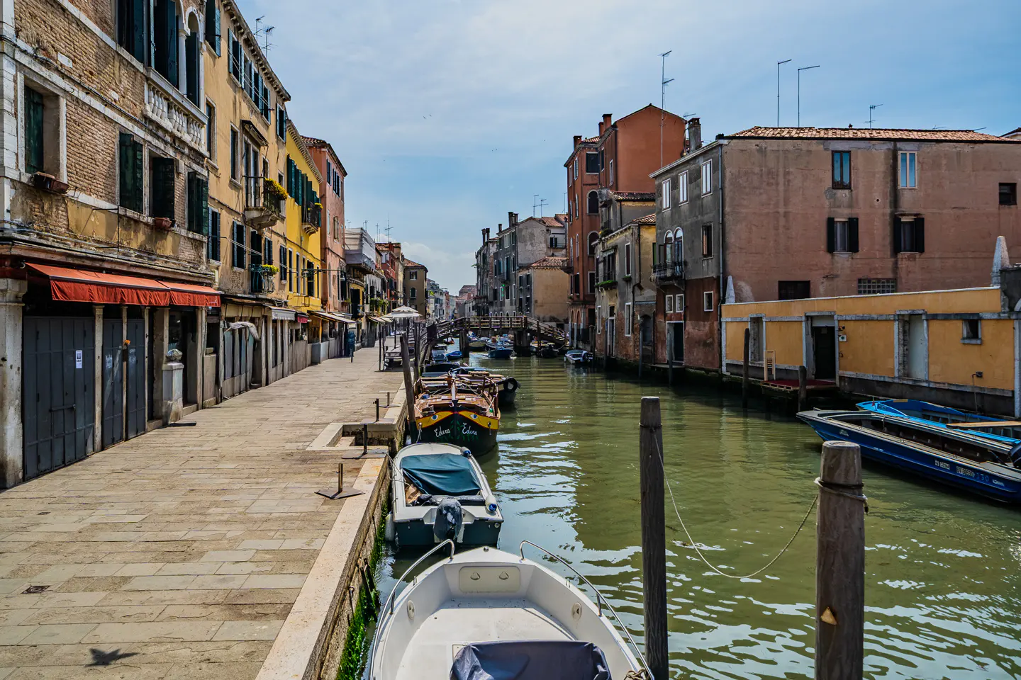 Venice canal view with boats docked along a stone walkway. Buildings line the canal, under a partly cloudy sky.