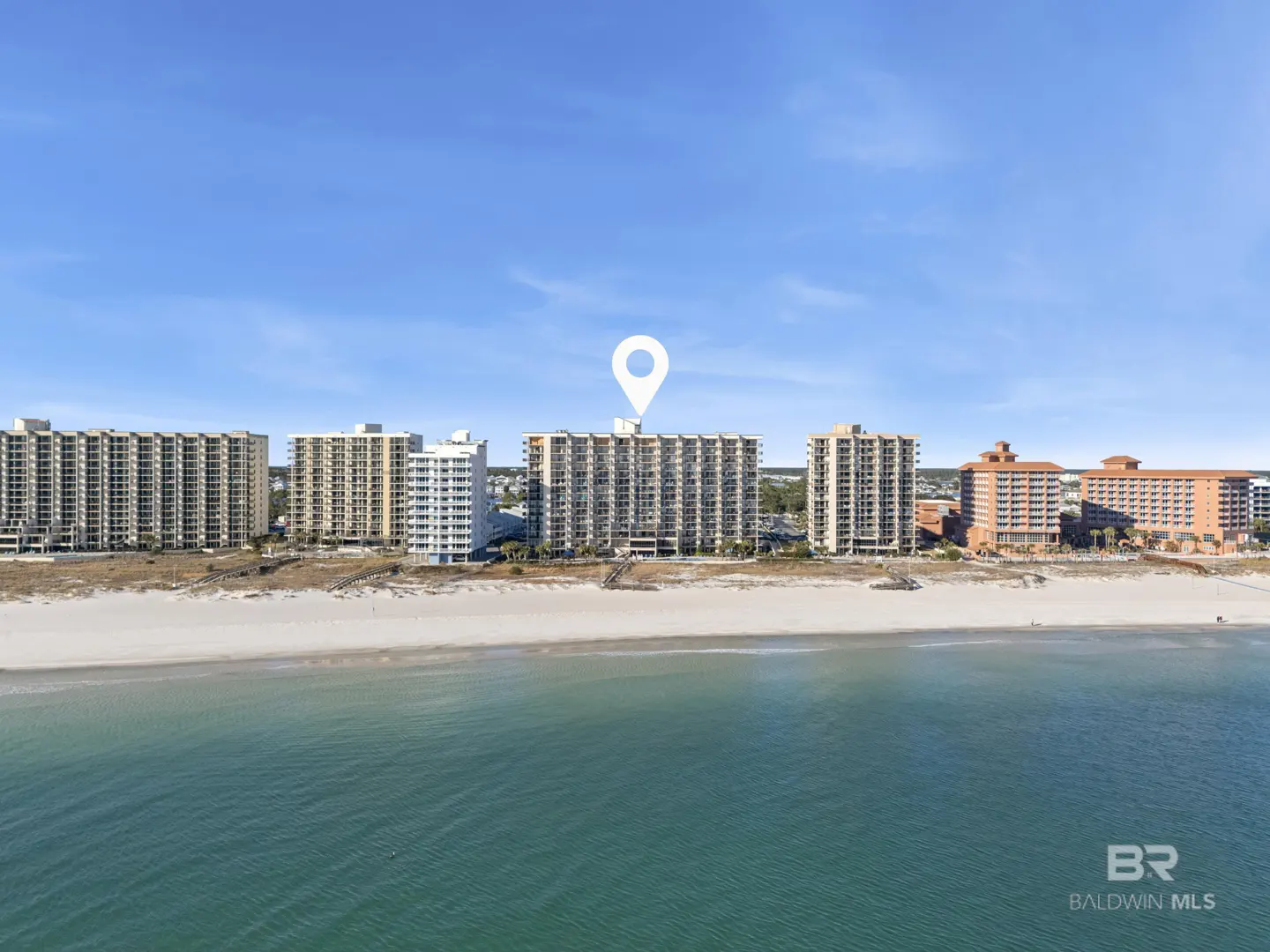 Aerial view of beachfront condos with a white location pin above the central building. Blue sky and ocean.