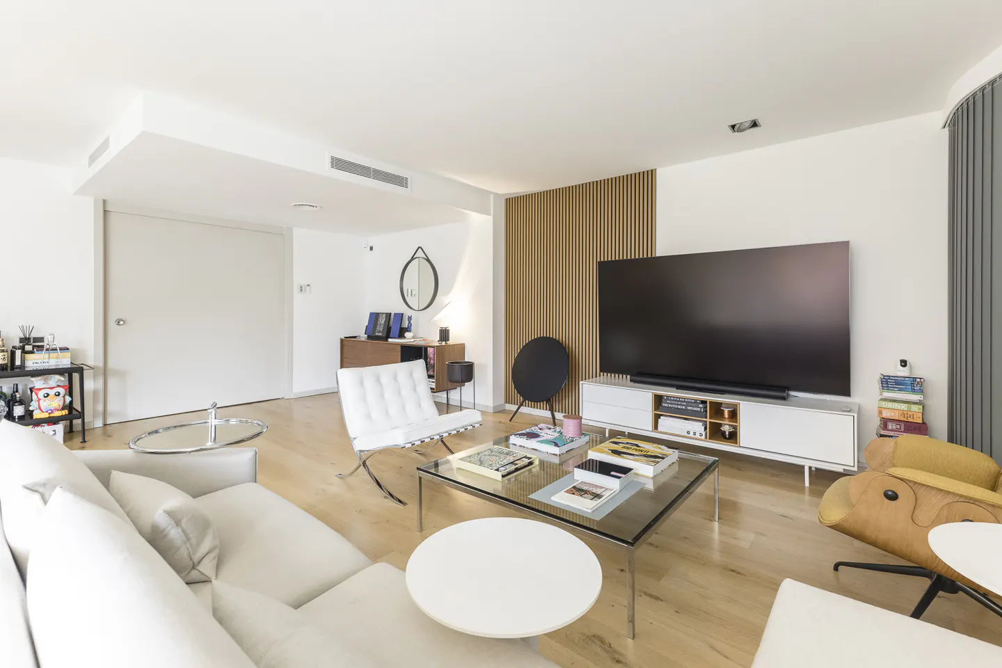 Modern living room with white sofa, Barcelona chair, glass coffee table, large TV on wood-slat wall, and light wood floors.