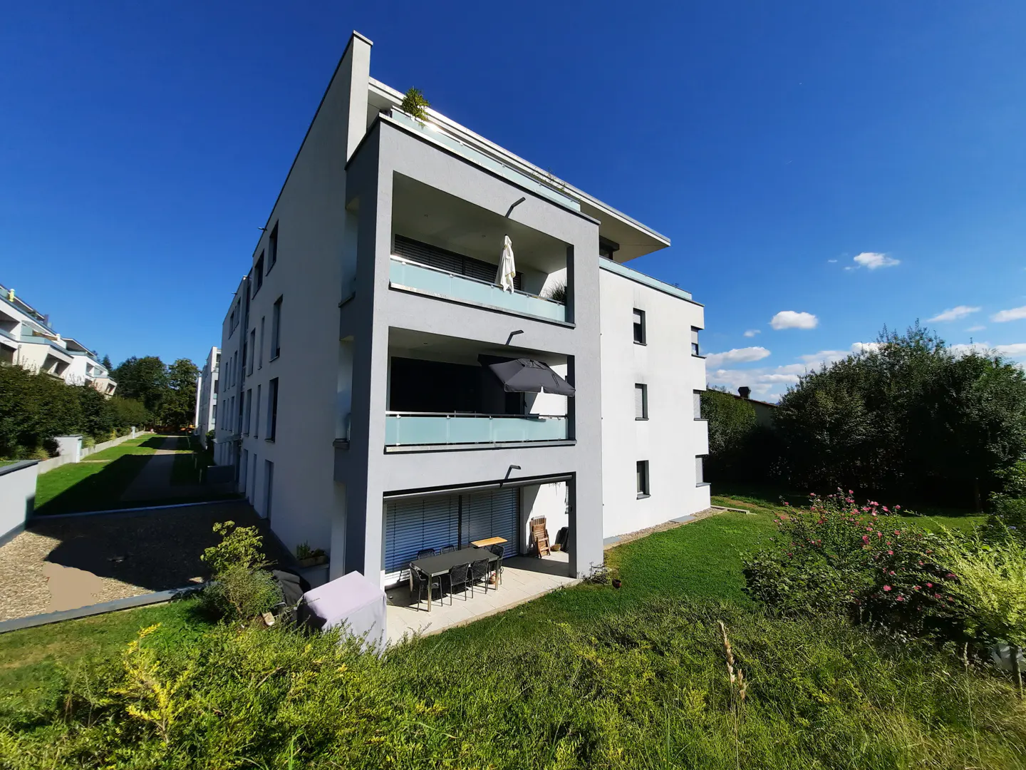 Modern white and gray apartment building with balconies, a patio with a table and chairs, and green grass under a blue sky.