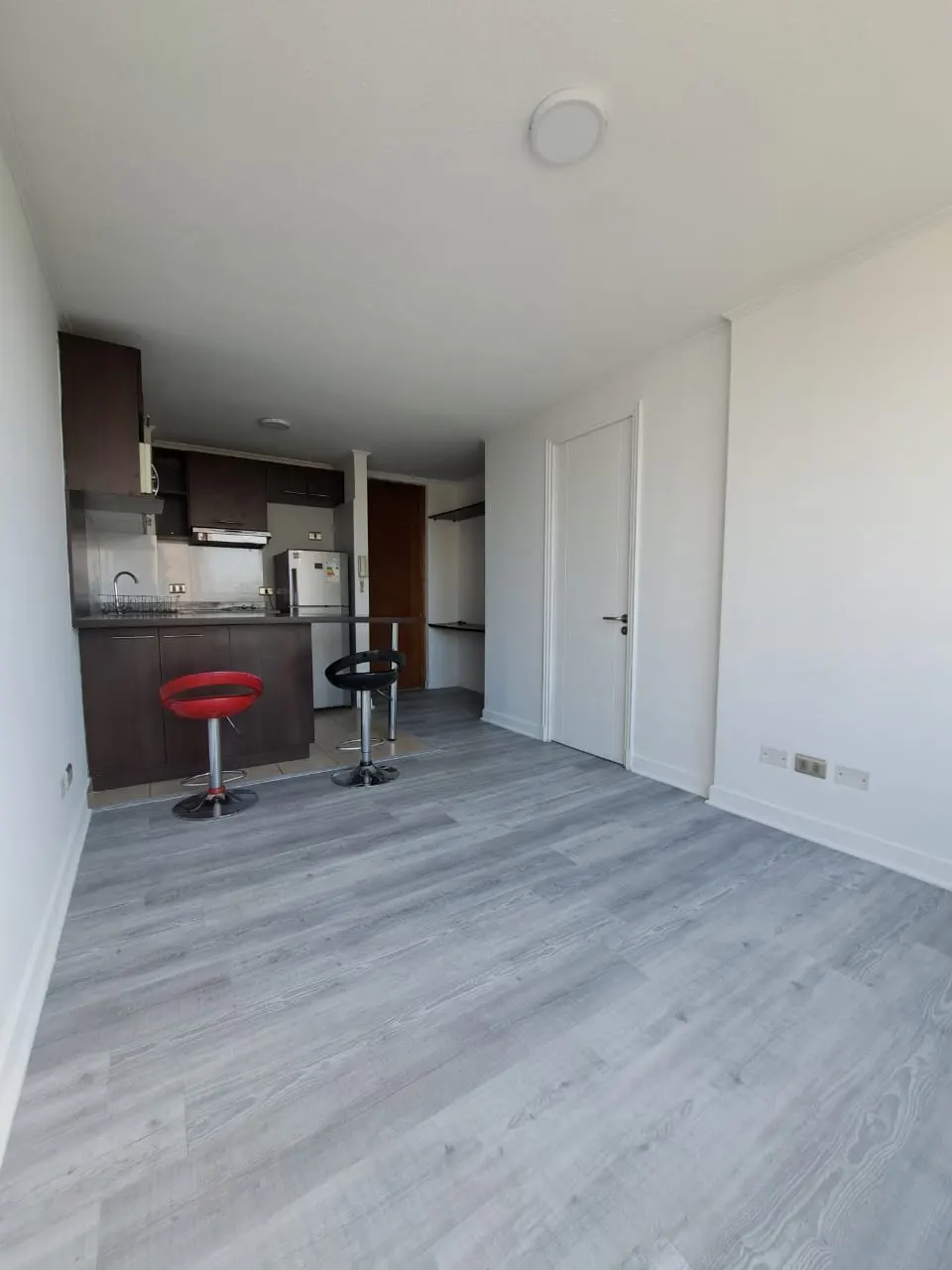 Open-concept apartment with gray wood floors, white walls, and a brown kitchen with red and black bar stools.